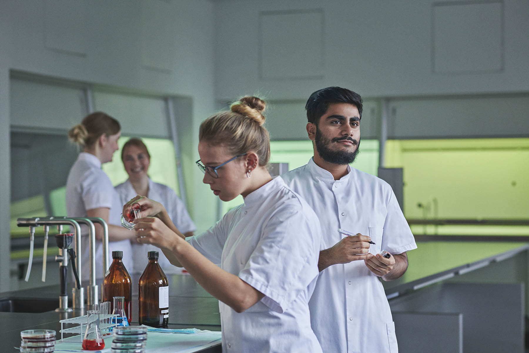 4 bioanalytikere-studerende på Københavns Professionshøjskole står i kitler i laberatorium og blander forskellige væsker.
