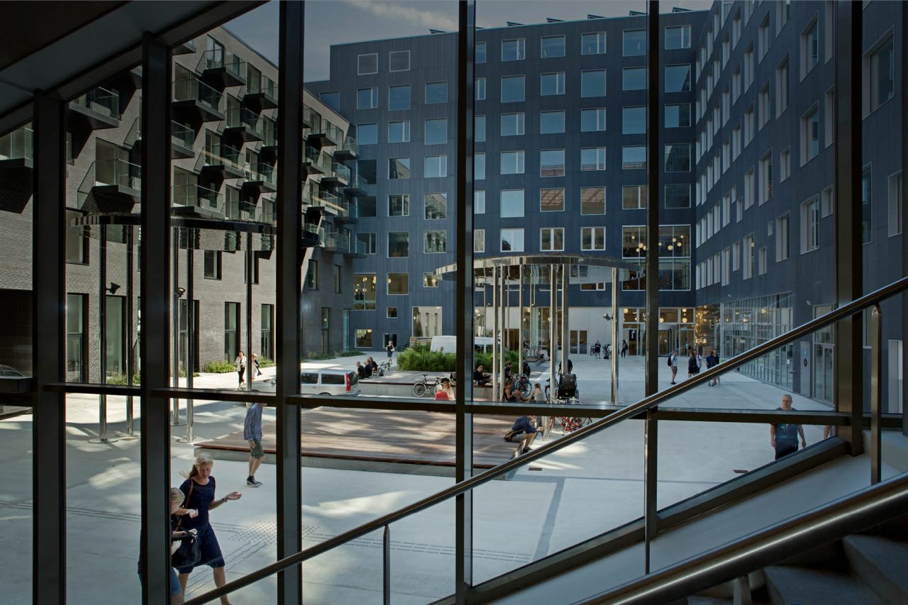 Der er mange mennesker og aktiviteter på torvet mellem bygningerne på campus carlsberg, som her ses gennem glasfacaden.