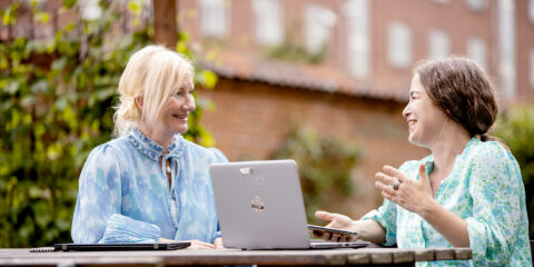 To personer sidder i en samtale udenfor på en bænk med hver deres bærbare computer og telefon