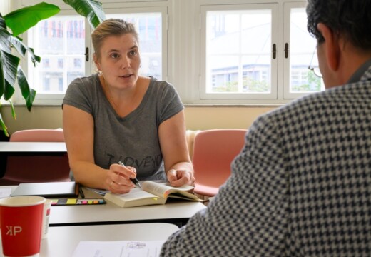 Undervisning i klasselokale, kvinde foran bog og i samtale med anden studerende foran sig.